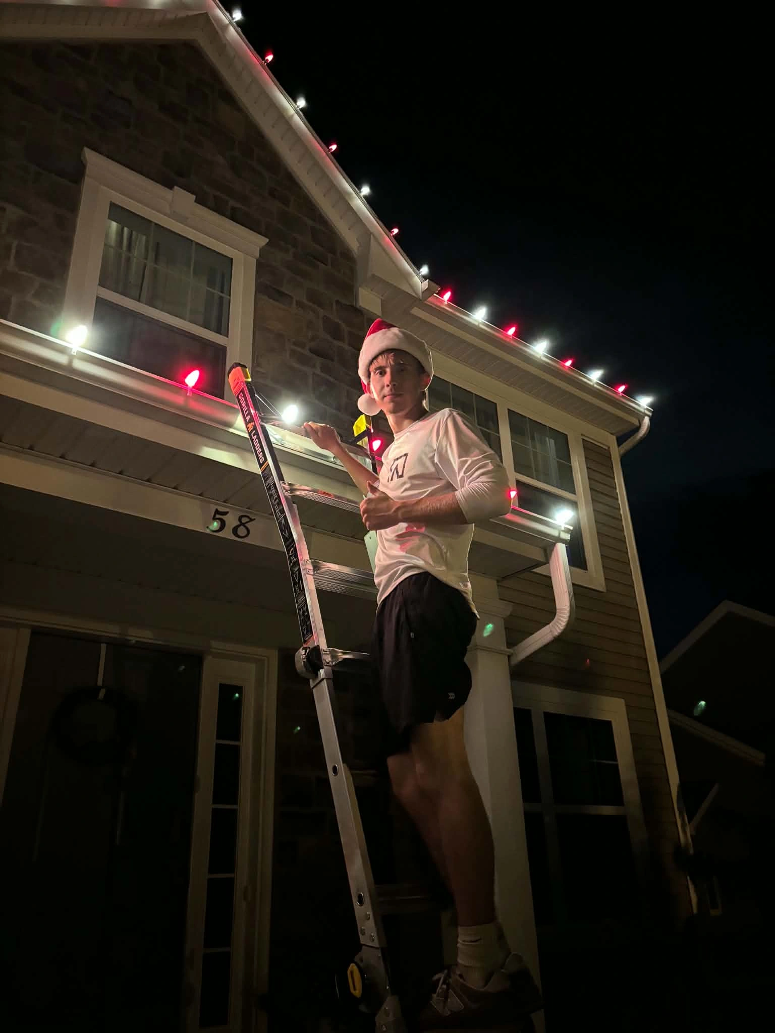 Christmas light installer wearing a Santa hat on a ladder beside red and white roofline lights